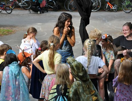 Kinder in Kostümen stehen im Kreis um eine Frau und einen Gitarrenspieler im Freien.