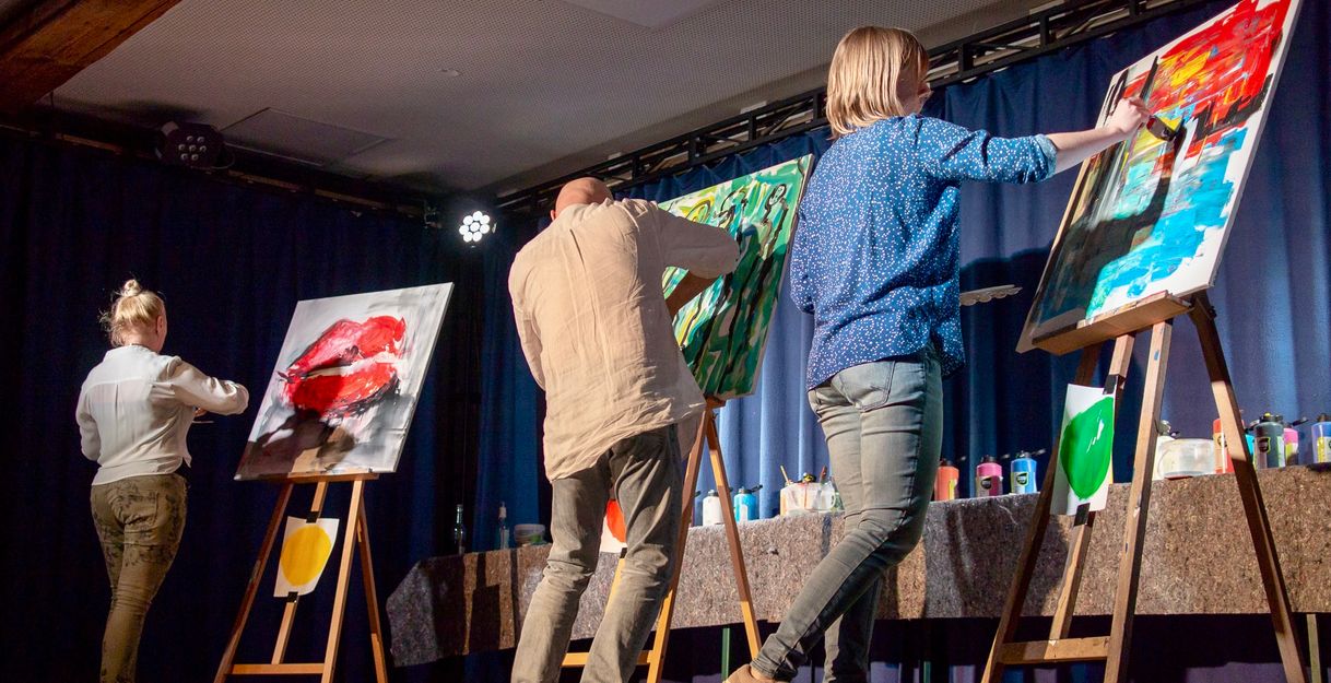 Auf dem Foto sind drei Künstler*innen auf der Bühne zu sehen, wie sie jeweils an einer Staffelei ihr Bild auf Leinwand malen.