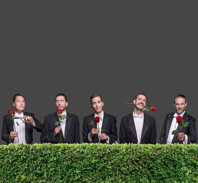 Fünf Männer in Anzügen mit Rosen hinter einer Hecke.