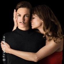 Zwei Frauen, eine mit aufgemaltem Schnurrbart, die andere umarmt sie von der Seite.