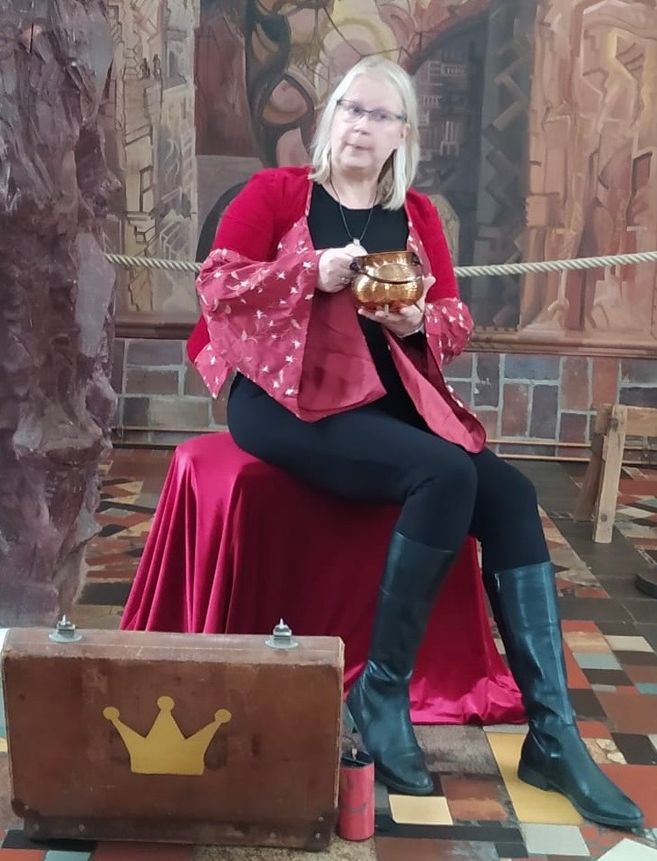 Eine Frau in rotem Gewand sitzt auf einem roten Stuhl und hält eine goldene Schale. Vor ihr steht ein Koffer mit einer Krone darauf.