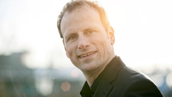 Porträt eines lächelnden Mannes im Anzug vor unscharfem Hintergrund mit Sonnenlicht.