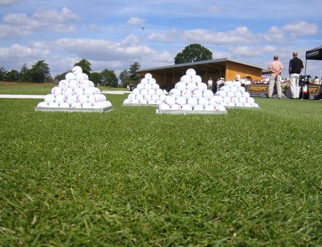 Golfbälle in Pyramidenform auf einer Driving Range mit Personen im Hintergrund.