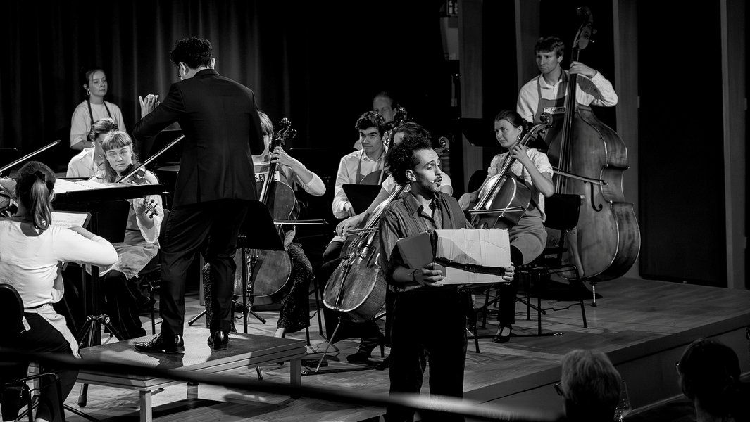 Ein Orchester spielt auf einer Bühne, während ein Mann mit einem Karton im Vordergrund steht.