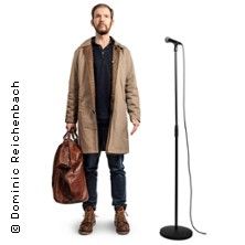 Ein Mann in einem beigen Mantel steht neben einem Mikrofonständer.