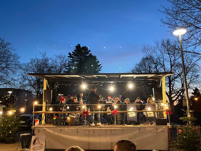 Eine Band spielt auf einer Bühne bei einem Weihnachtsmarkt in Hemmoor, umgeben von Lichtern und Weihnachtsbäumen.