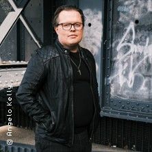 Person in schwarzer Lederjacke vor einer Wand mit Graffiti.