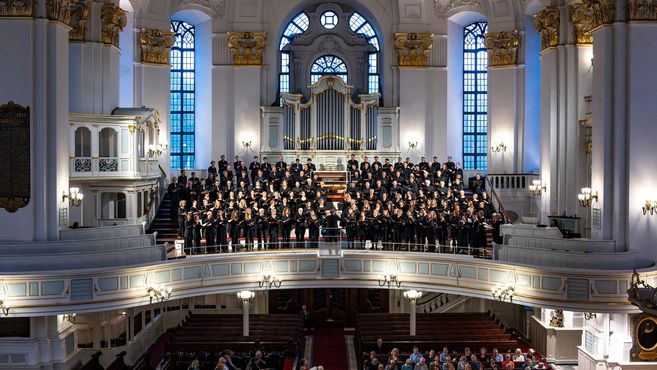 Ein großer Chor singt auf einer Empore in einer prächtigen Kirche mit Orgel im Hintergrund.