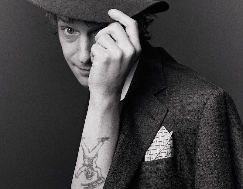 Schwarz-weiß Foto eines Mannes mit Hut, der seinen Arm hebt und ein Tattoo zeigt.