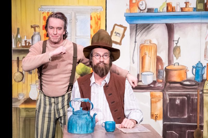 Zwei Schauspieler in einem Bühnenbild, das eine Küche darstellt. Einer sitzt mit Hut und Bart, der andere steht daneben.