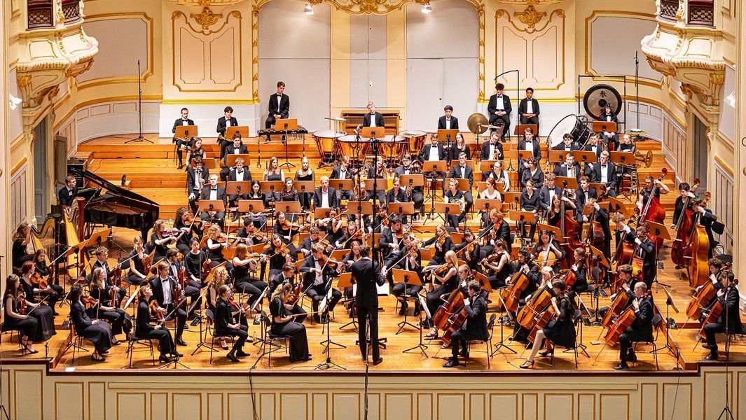 Ein großes Orchester spielt auf einer Bühne in einem Konzertsaal.