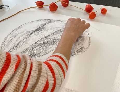 eine Frau in einem rot-weiß geringeltem Pullover zeichnet mit Kohle vergrößert einen Stengel mit orangenen Lampionblumen 
