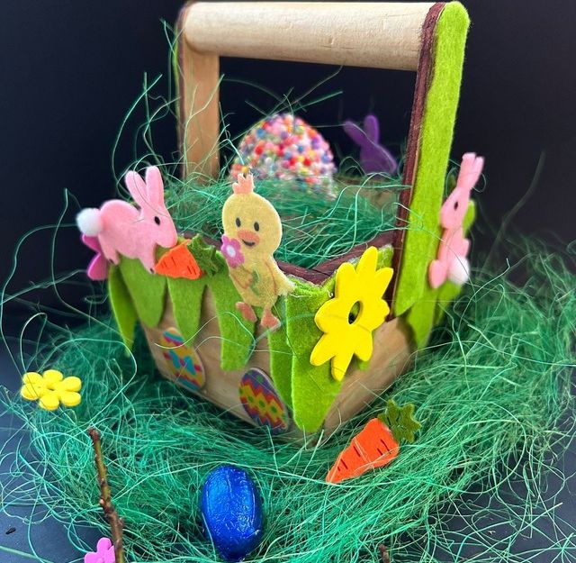 Ein dekorativer Osterkorb mit Filzhasen, Küken und Blumen, umgeben von grünem Kunstgras und einem blauen Osterei.