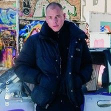 Person in schwarzer Jacke vor einem Auto mit Graffiti im Hintergrund.