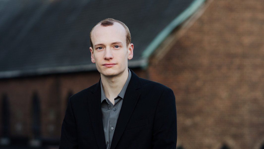 Ein junger Mann in einem schwarzen Anzug steht vor einem unscharfen Hintergrund mit Backsteinmauer.