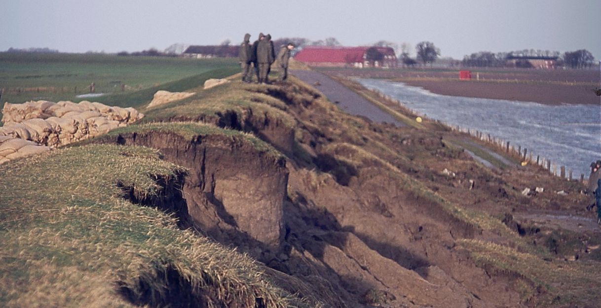 Erosion an einem Deich nach einer Sturmflut, mit Sandsäcken und Menschen im Hintergrund.