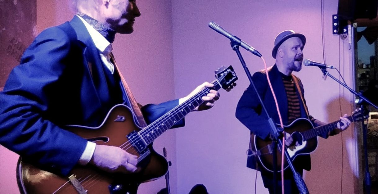 zwei Gitarristen, einer links im Vordergrund, der andere rechts im Hintergund spielen vor ihren Mirkrofonen