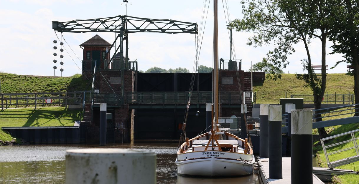 Ein Segelboot liegt vor der Schleuse Kasenort, umgeben von grünen Wiesen und Bäumen.