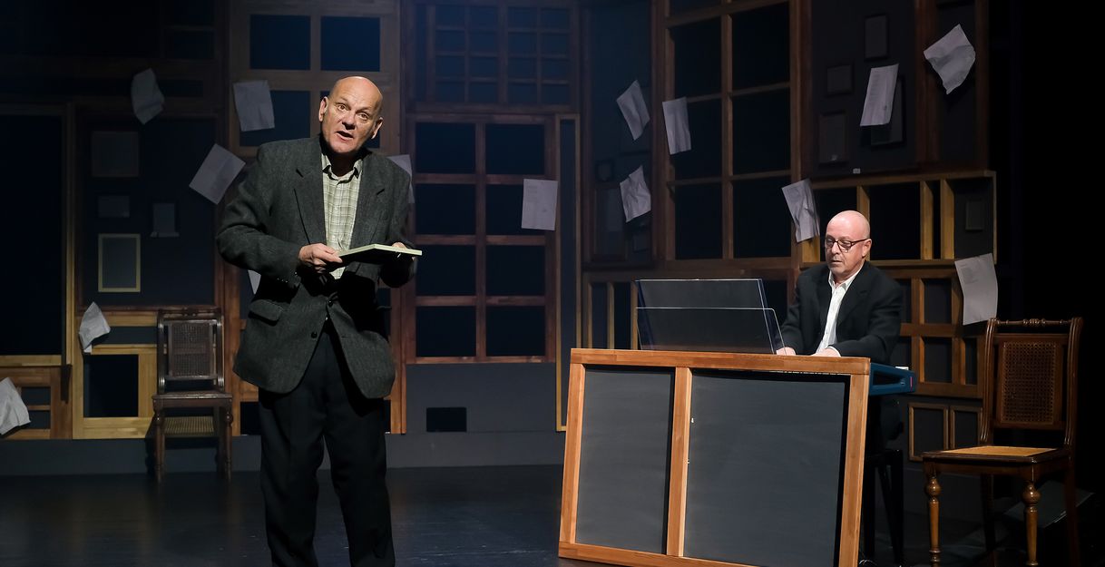 Zwei Männer auf einer Bühne, einer liest aus einem Buch, der andere spielt Klavier. Der Hintergrund ist mit Papierblättern dekoriert.