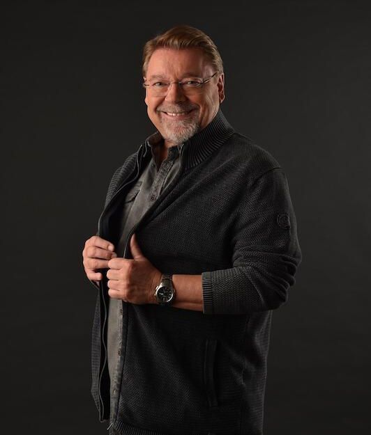 Ein lächelnder Mann in dunkler Kleidung vor einem schwarzen Hintergrund.