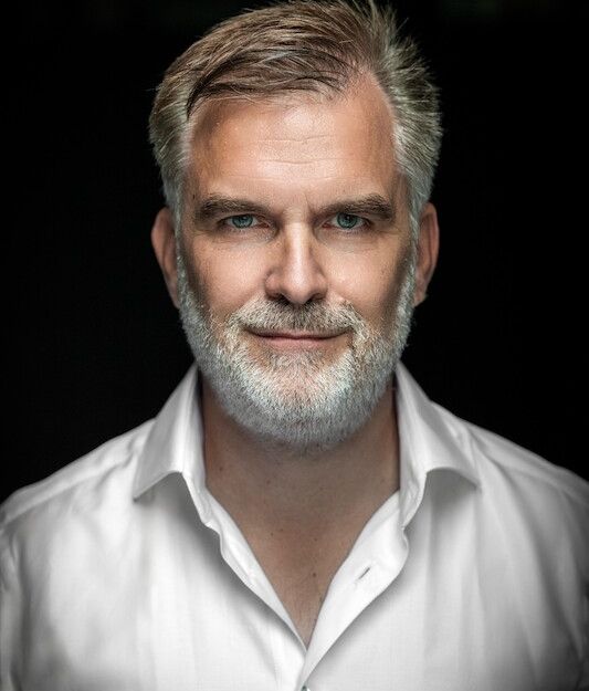 Porträt eines Mannes mit grauem Bart und weißem Hemd vor dunklem Hintergrund.