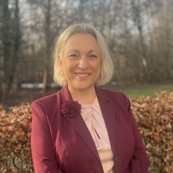 Eine lächelnde Frau in einem bordeauxfarbenen Blazer steht vor einer Hecke im Freien.