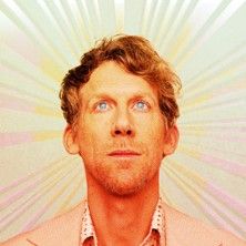 Porträt eines Mannes mit blondem Haar und blauem Hemd vor einem strahlenförmigen Hintergrund.