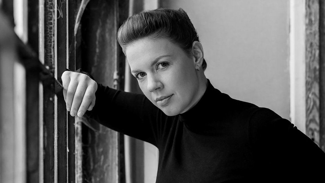 Schwarz-weiß Porträt einer Frau in einem schwarzen Rollkragenpullover, die an einem Fensterrahmen lehnt.