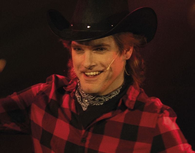 Person mit Cowboyhut und kariertem Hemd auf einer Bühne.