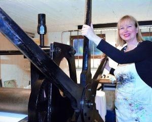 Eine Frau steht neben einer großen Druckerpresse in einem Atelier.