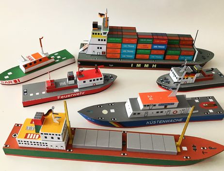 Papiermodelle von verschiedenen Schiffen, darunter ein Containerschiff, ein Küstenwachboot und ein Feuerwehrboot, auf einem Tisch arrangiert.
