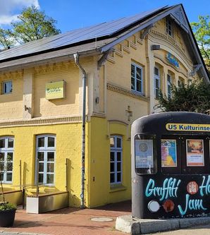 Gelbes Backsteingebäude mit Schildern von Brakula und Vistro, daneben eine bemalte U-Bahn-Attrappe mit Graffiti.