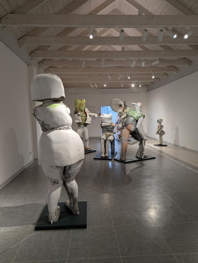 A piece of art in an exhibition space with several sculptural figures. The figures have different colors and sizes and stand on black pedestals.