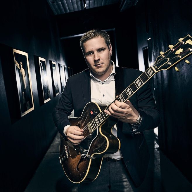 Ein Mann in einem Anzug spielt Gitarre in einem dunklen Flur mit gerahmten Bildern an den Wänden.