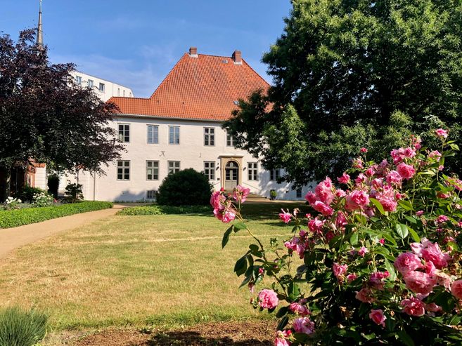 Ein historisches Gebäude mit rotem Ziegeldach, umgeben von einem gepflegten Garten mit blühenden Rosen im Vordergrund.