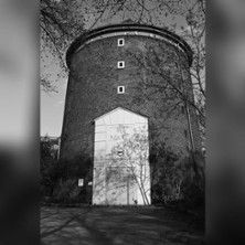 Schwarzweißfoto eines runden Zombeck-Turms mit hoher Eingangstür und kleinen Fenstern, umgeben von Bäumen.
