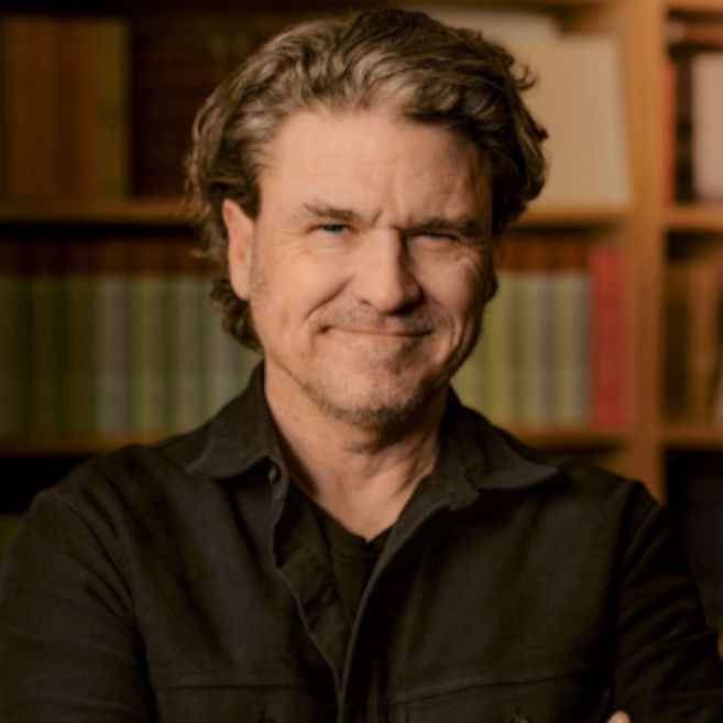 Ein Mann mit grauen Haaren und Bart lächelt vor einem Bücherregal.