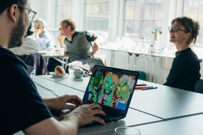 Person arbeitet an einem Laptop mit einer Zeichnung von Cartoon-Zombies, während andere im Hintergrund an einem Tisch sitzen und diskutieren.