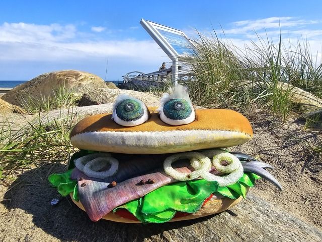 Ein großes Fischbrötchen-Kostüm mit Augen liegt auf einem Felsen am Strand, im Hintergrund eine Seebrücke und das Meer.