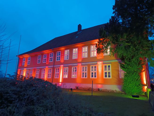 Ein beleuchtetes Gebäude bei Dämmerung, mit roten und orangen Lichtern angestrahlt.