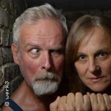 Zwei Personen stehen vor einer Steinwand, eine mit ernstem Gesichtsausdruck, die andere mit verschränkten Händen.