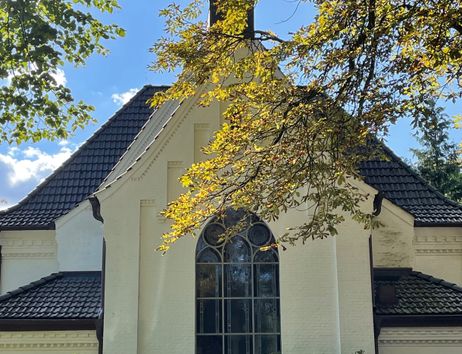 Eine weiße Kirche mit einem großen Fenster und einem schwarzen Dach, umgeben von Bäumen und Sonnenlicht.