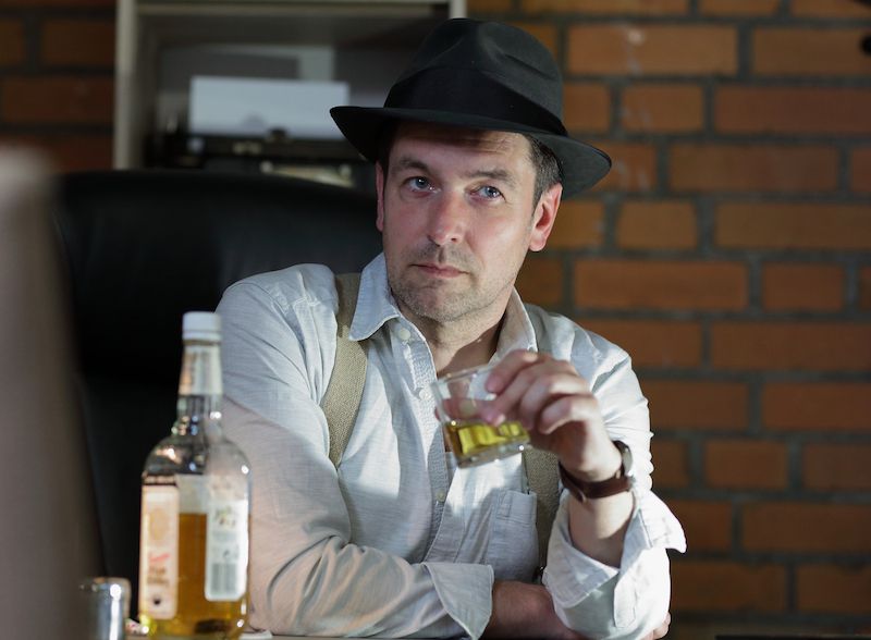 Mann mit Hut und Glas in der Hand, sitzt vor einer Ziegelwand.