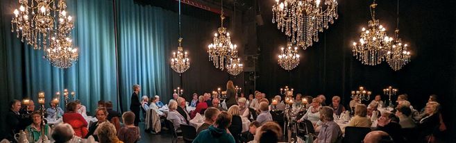 Menschen sitzen an gedeckten Tischen in einem Theatersaal mit Kronleuchtern.