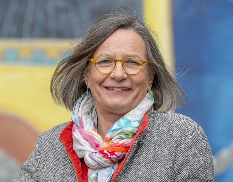 Eine Frau mit grauem Haar und Brille lächelt in die Kamera. Sie trägt einen bunten Schal und einen grauen Mantel. Der Hintergrund ist unscharf und farbenfroh.