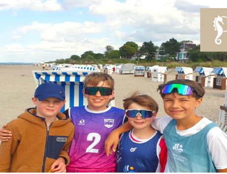 Vier Kinder stehen am Strand vor Strandkörben und lächeln in die Kamera. Sie tragen sportliche Kleidung und Sonnenbrillen.