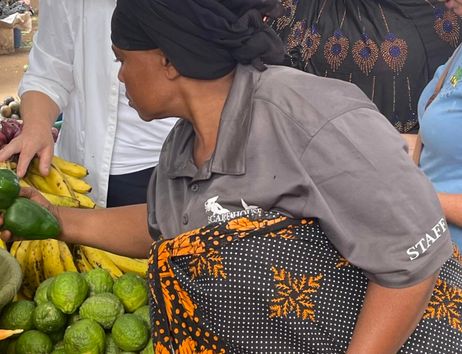 In the picture, people can be seen at a market selling and buying fresh fruits and vegetables. The atmosphere is lively and colorful, and the vendors are wearing traditional clothing.