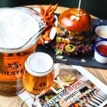 Ein Tisch mit einem Bierkrug, einem Glas Bier, einem Burger mit Pommes und einem Super Bowl LX Flyer.