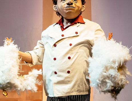 Ein Schauspieler in einem weißen Kochkostüm mit einer großen Maske hält zwei Hühner aus Federn in den Händen.