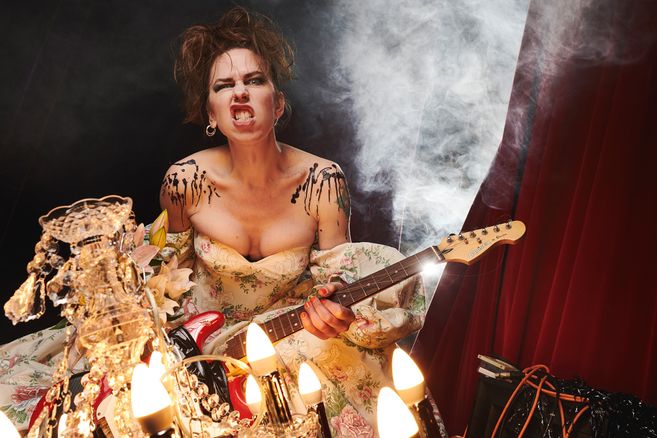 Emma Frankland, eine weiblich gelesene Person mit braunen Haaren steht mit einer E-Gitarre und rockigem Gesichtsausdruck vor einem Vorhang. Im Hintergrund ist Rauch zu sehen, während im Vordergrund ein Kronleuchter ist.